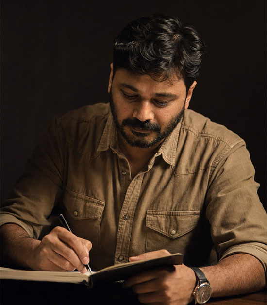 Man sitting in a dimly lit room, wearing a beige shirt and looking contemplative.